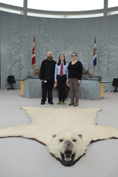 Michael Ball, Manager, Journals, Kagan Ball (Youth Parliamentarian MLA Yellowknife South), and Dana Ball infront of Mace and Speaker's Chair. Polar bear rug in foreground.