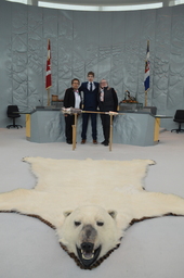 Northwest Territories Commissioner, Margaret Thom, Hunter Groenewegen (Youth Parliamentarian MLA Hay River North) and Jane Groenewegan (grandmother) former Member of the Northwest Territories Legislative Assembly standing behind the Mace. Polar bear rug in the foreground.