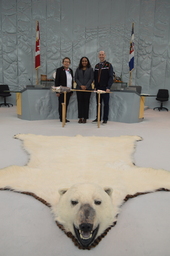 Northwest Territories Commissioner, Margaret Thom, Ritaj Hamad-Dawoud (Youth Parliamentarian MLA Yellowknife Centre) and Robert Hawkins, MLA Yellowknife Centre standing behind the Mace. Polar bear rug in the foreground.